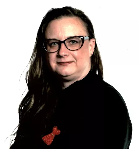 A woman with long hair and glasses, wearing a black shirt with a beaded red dress pin.